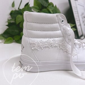 K&ouml;nnte beinhalten: Ein wei&szlig;er High-Top-Sneaker mit gepolstertem Kn&ouml;chelbereich, wei&szlig;en Satin-Schn&uuml;rsenkeln und einer feinen wei&szlig;en Spitzenverzierung an der Seite. Im Fersenbereich ist ein Teil eines schwarzen Schriftzugs zu sehen, die Sohle ist aus wei&szlig;em Gummi.