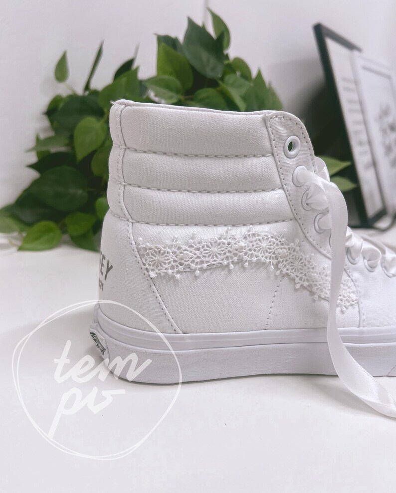 K&ouml;nnte beinhalten: Ein wei&szlig;er High-Top-Sneaker mit gepolstertem Kn&ouml;chelbereich, wei&szlig;en Satin-Schn&uuml;rsenkeln und einer feinen wei&szlig;en Spitzenverzierung an der Seite. Im Fersenbereich ist ein Teil eines schwarzen Schriftzugs zu sehen, die Sohle ist aus wei&szlig;em Gummi.