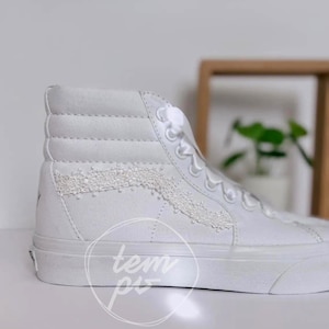 K&ouml;nnte beinhalten: Ein hoher Sneaker aus wei&szlig;em Canvas mit einer dekorativen wei&szlig;en Spitzenborte an der Seitenpartie. Der Schuh verf&uuml;gt &uuml;ber wei&szlig;e Schn&uuml;rsenkel, einen gepolsterten Schaft und eine dicke wei&szlig;e Gummisohle. Ein kreisf&ouml;rmiges Wasserzeichen mit tem pi ist sichtbar.