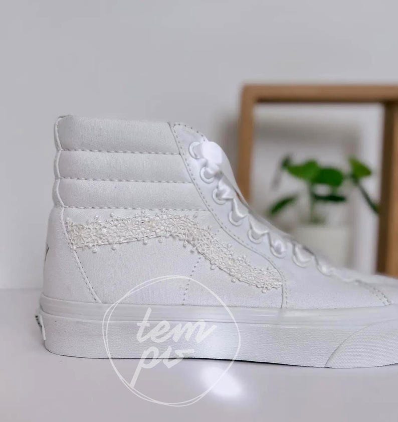 K&ouml;nnte beinhalten: Ein hoher Sneaker aus wei&szlig;em Canvas mit einer dekorativen wei&szlig;en Spitzenborte an der Seitenpartie. Der Schuh verf&uuml;gt &uuml;ber wei&szlig;e Schn&uuml;rsenkel, einen gepolsterten Schaft und eine dicke wei&szlig;e Gummisohle. Ein kreisf&ouml;rmiges Wasserzeichen mit tem pi ist sichtbar.