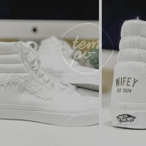 K&ouml;nnte beinhalten: Ein Paar wei&szlig;e High-Top-Sneaker mit Spitzenbesatz an den Seitenteilen und wei&szlig;en Schn&uuml;rsenkeln. Die Ferse des Schuhs zeigt den Text WIFEY EST 2024 in dunkler Schrift sowie ein Vans Off The Wall-Logo auf der Gummisohle an der Ferse.