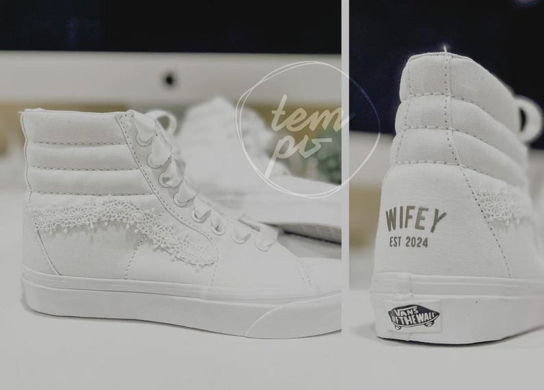 K&ouml;nnte beinhalten: Ein Paar wei&szlig;e High-Top-Sneaker mit Spitzenbesatz an den Seitenteilen und wei&szlig;en Schn&uuml;rsenkeln. Die Ferse des Schuhs zeigt den Text WIFEY EST 2024 in dunkler Schrift sowie ein Vans Off The Wall-Logo auf der Gummisohle an der Ferse.
