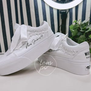 K&ouml;nnte beinhalten: Ein Paar wei&szlig;e Plateau-Sneaker mit breiten Satin-Schn&uuml;rsenkeln, zarter wei&szlig;er Spitzenborte an den Seitenteilen und individueller schwarzer Beschriftung, die Mrs. Jenkins mit einem Herz sowie WIFE 10.10.23 an der Ferse zeigt.