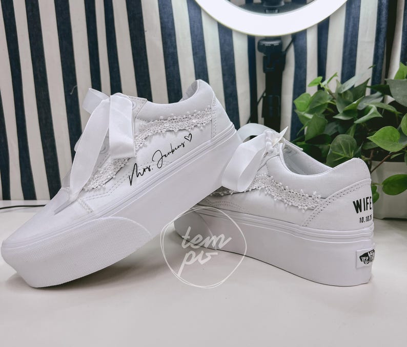 K&ouml;nnte beinhalten: Ein Paar wei&szlig;e Plateau-Sneaker mit breiten Satin-Schn&uuml;rsenkeln, zarter wei&szlig;er Spitzenborte an den Seitenteilen und individueller schwarzer Beschriftung, die Mrs. Jenkins mit einem Herz sowie WIFE 10.10.23 an der Ferse zeigt.