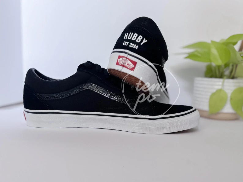 May include: Black low-top trainers with a white rubber sole and a black side stripe. The heel features the white text HUBBY EST 2024 above a red and white Vans Off The Wall logo patch. The canvas upper has a textured finish along the side panel.