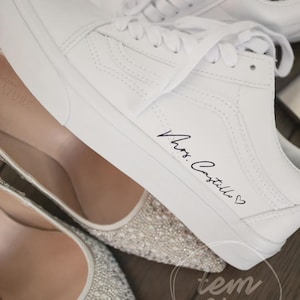 Puede incluir: Zapatillas de lona blancas con cordones, con la inscripción "Mrs. Castillo" en escritura negra con un símbolo de corazón. La imagen también muestra un par de tacones adornados con punta.