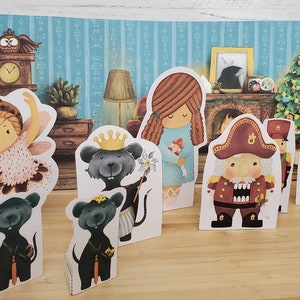 May include: A set of paper doll characters from the Nutcracker story. The characters include a ballerina, a nutcracker, a mouse king, a girl, and two toy soldiers. The dolls are standing on a wooden surface in front of a backdrop of a Christmas tree and a fireplace.