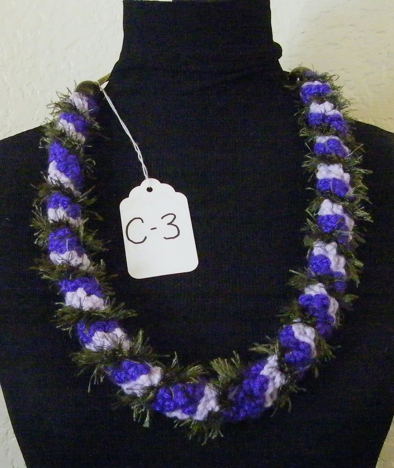 May include: Purple and white crocheted lei with green accents. The lei is displayed on a black mannequin. A white tag with the text "C-3" is attached to the lei.