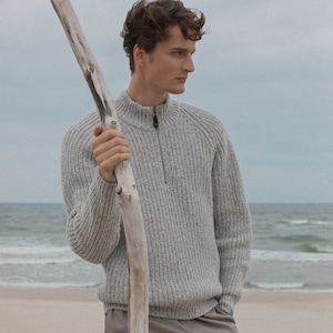 May include: A light gray ribbed knit sweater with a quarter-zip collar. The model is standing on a sandy beach, holding a piece of driftwood. The sweater has a high neckline and long sleeves. The pants are a light brown color.