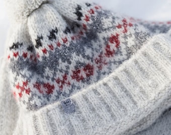 May include: A white knit beanie with a pom pom on top and a red, gray, and black patterned design. The beanie is folded over and has a white fringe.