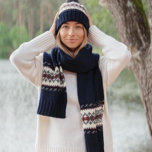 May include: A woman wearing a white sweater, white pants, a blue and white knit hat, and a blue and white knit scarf. She is standing in front of a lake and trees.