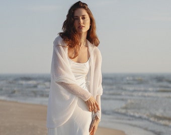 May include: A woman wearing a white dress and a white cardigan stands on a sandy beach. The ocean is in the background.