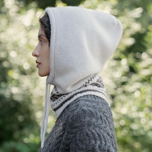 Peut inclure: Un capuchon en tricot gris clair avec un cordon de serrage, associé à un pull gris assorti. Le capuchon présente un motif autour du cou. Le pull a un motif en tricot torsadé texturé.