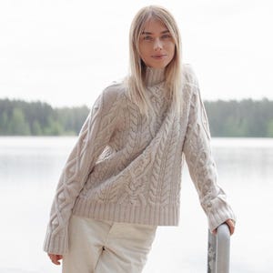 May include: A woman wearing a light beige cable knit sweater with a turtleneck. The sweater has a loose fit and a slightly cropped length. The woman is standing in front of a lake with trees in the background.