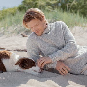 Puede incluir: Un hombre con un su&eacute;ter gris claro se sienta en una playa de arena con un perro marr&oacute;n y blanco. El hombre est&aacute; acariciando al perro, que est&aacute; acostado de lado.