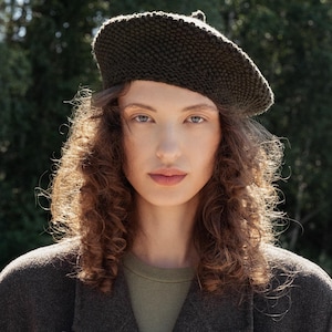 May include: A woman wearing a dark green knit beret and a dark brown coat. The beret is made of a textured knit material. The coat has a classic double-breasted design. The woman is also wearing a green shirt.
