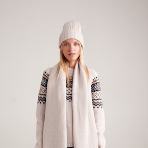 May include: A beige ribbed knit beanie, a long beige knit scarf, and a beige long-sleeve sweater featuring a brown, blue, and white geometric pattern across the chest and shoulders, paired with dark blue denim jeans.
