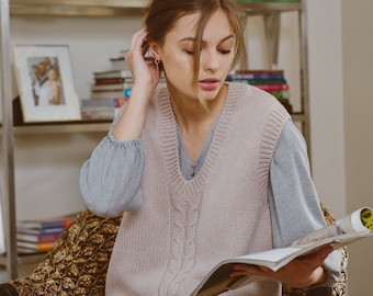 May include: A woman wearing a light pink cable knit sweater vest over a gray long-sleeved shirt, sitting in a wicker chair and reading a magazine.