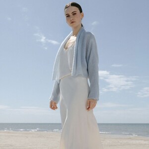 May include: A light blue open-front knit cardigan with long sleeves and a soft texture, worn over a simple white sleeveless dress. The cardigan features a relaxed fit and a draped collar, providing a lightweight layer over the smooth white dress.