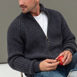 May include: A dark gray knitted zip-up cardigan with a white t-shirt underneath. The person is holding a red apple.