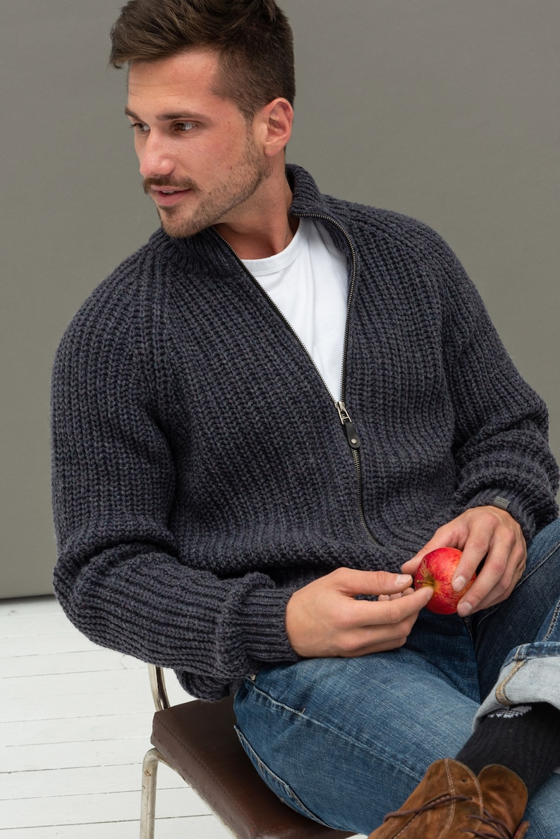 May include: A dark gray knitted zip-up cardigan with a white t-shirt underneath. The person is holding a red apple.