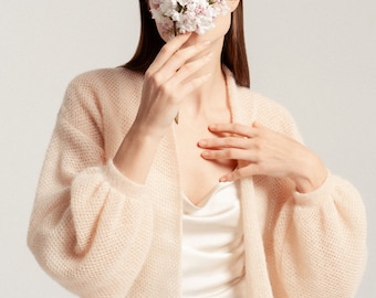 May include: A pale peach, open-front knit cardigan featuring a textured, airy weave and voluminous balloon sleeves. The garment is worn over a smooth, cream-coloured satin slip dress. A cluster of light pink and white lilac flowers is held in front.