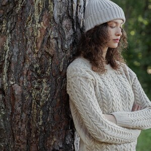 May include: A beige cable-knit sweater with a crew neckline and long sleeves, paired with a matching beige ribbed knit beanie. The sweater features a vertical braided pattern throughout the front and sleeves, with ribbed cuffs and a ribbed hem.