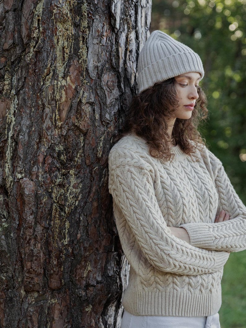 May include: A beige cable-knit sweater with a crew neckline and long sleeves, paired with a matching beige ribbed knit beanie. The sweater features a vertical braided pattern throughout the front and sleeves, with ribbed cuffs and a ribbed hem.