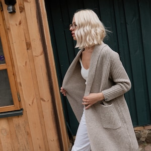 Pu&ograve; includere: Una donna indossa un cardigan marrone chiaro con una canottiera bianca e pantaloni bianchi. Sta camminando per strada con un edificio in legno e un muro verde sullo sfondo.