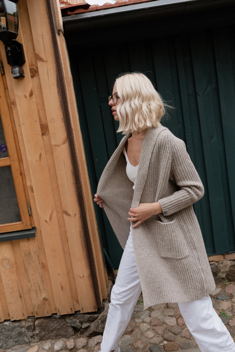 Pu&ograve; includere: Una donna indossa un cardigan marrone chiaro con una canottiera bianca e pantaloni bianchi. Sta camminando per strada con un edificio in legno e un muro verde sullo sfondo.