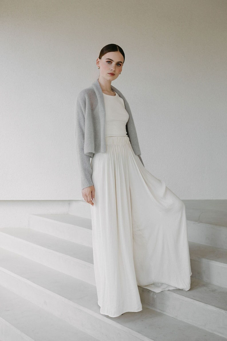 May include: A light gray open-front knit cardigan worn over a cream-colored sleeveless top and high-waisted, floor-length wide-leg trousers with a gathered elastic waistband and a flowing, pleated silhouette.