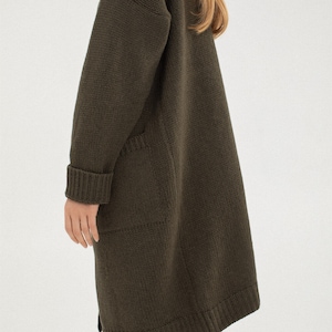 May include: A long, dark green knitted cardigan with a relaxed fit and two pockets. The cardigan is made of a soft, comfortable material and has a ribbed texture.