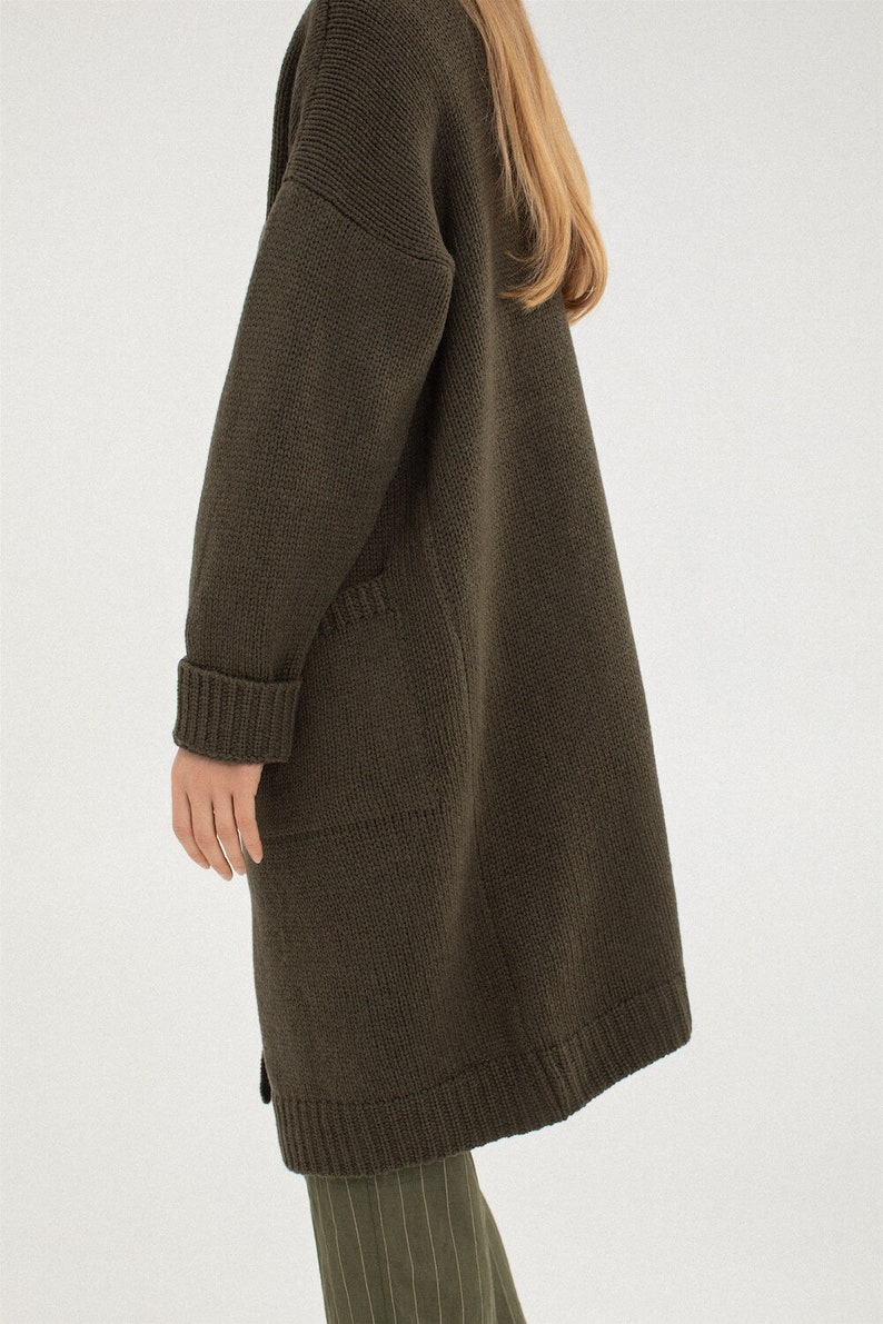 May include: A long, dark green knitted cardigan with a relaxed fit and two pockets. The cardigan is made of a soft, comfortable material and has a ribbed texture.
