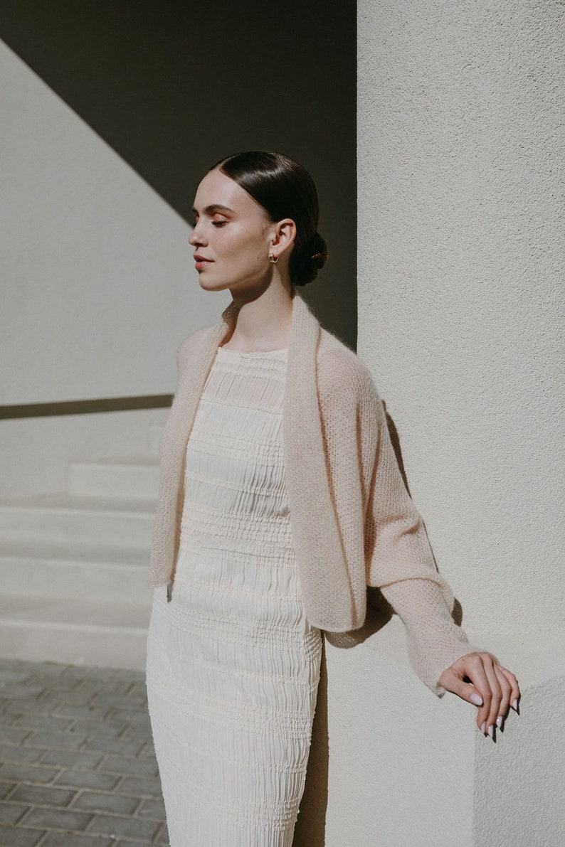May include: A cream-colored, long-sleeved, open-front knit shrug made of a soft, sheer material, worn over a sleeveless, off-white, textured, smocked-bodice dress that features a high neckline and a form-fitting, knee-length silhouette.