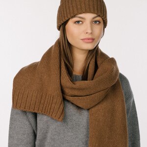 May include: A brown ribbed knit beanie with a folded cuff and a matching long brown ribbed knit scarf. The beanie and scarf are made of a solid brown yarn and feature a consistent vertical ribbed texture throughout the entire design.