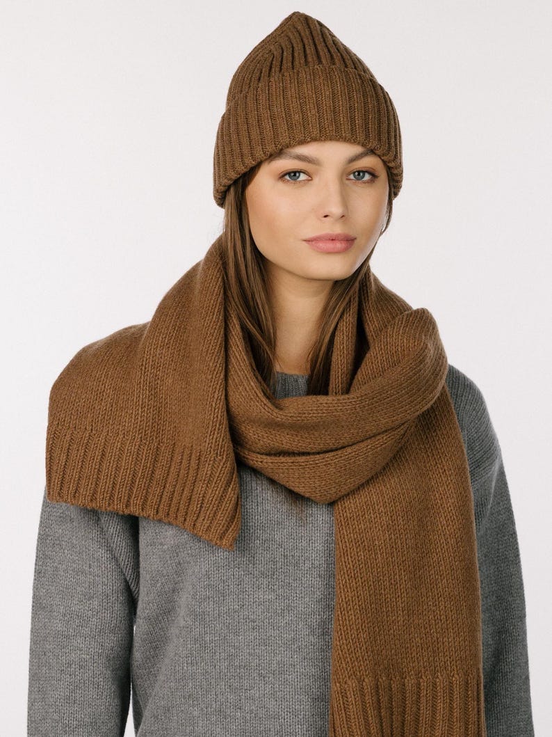 May include: A brown ribbed knit beanie with a folded cuff and a matching long brown ribbed knit scarf. The beanie and scarf are made of a solid brown yarn and feature a consistent vertical ribbed texture throughout the entire design.