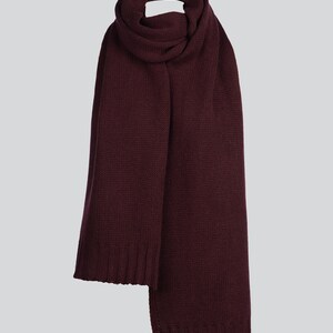 May include: A deep burgundy knit scarf featuring a consistent textured stitch pattern throughout the length of the fabric. The scarf has ribbed edges at both ends, providing a structured finish to the soft, solid-colored winter accessory.