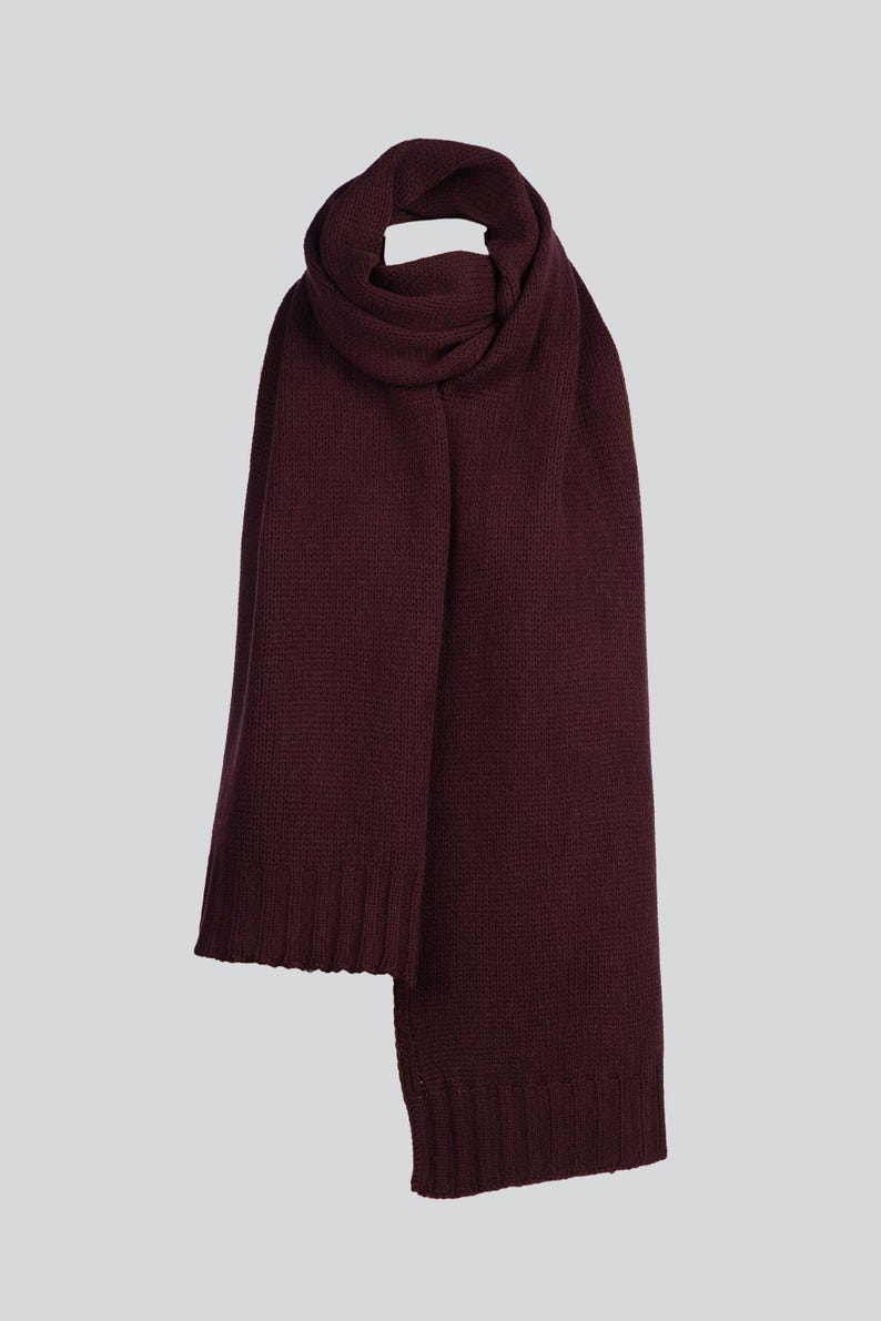 May include: A deep burgundy knit scarf featuring a consistent textured stitch pattern throughout the length of the fabric. The scarf has ribbed edges at both ends, providing a structured finish to the soft, solid-colored winter accessory.