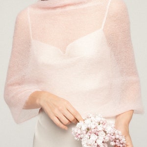 May include: A pale pink, sheer, knitted shawl with a wide boat neckline and long sleeves, worn over a white camisole. The garment features a delicate, airy texture and a loose, draped fit that covers the shoulders and upper arms.