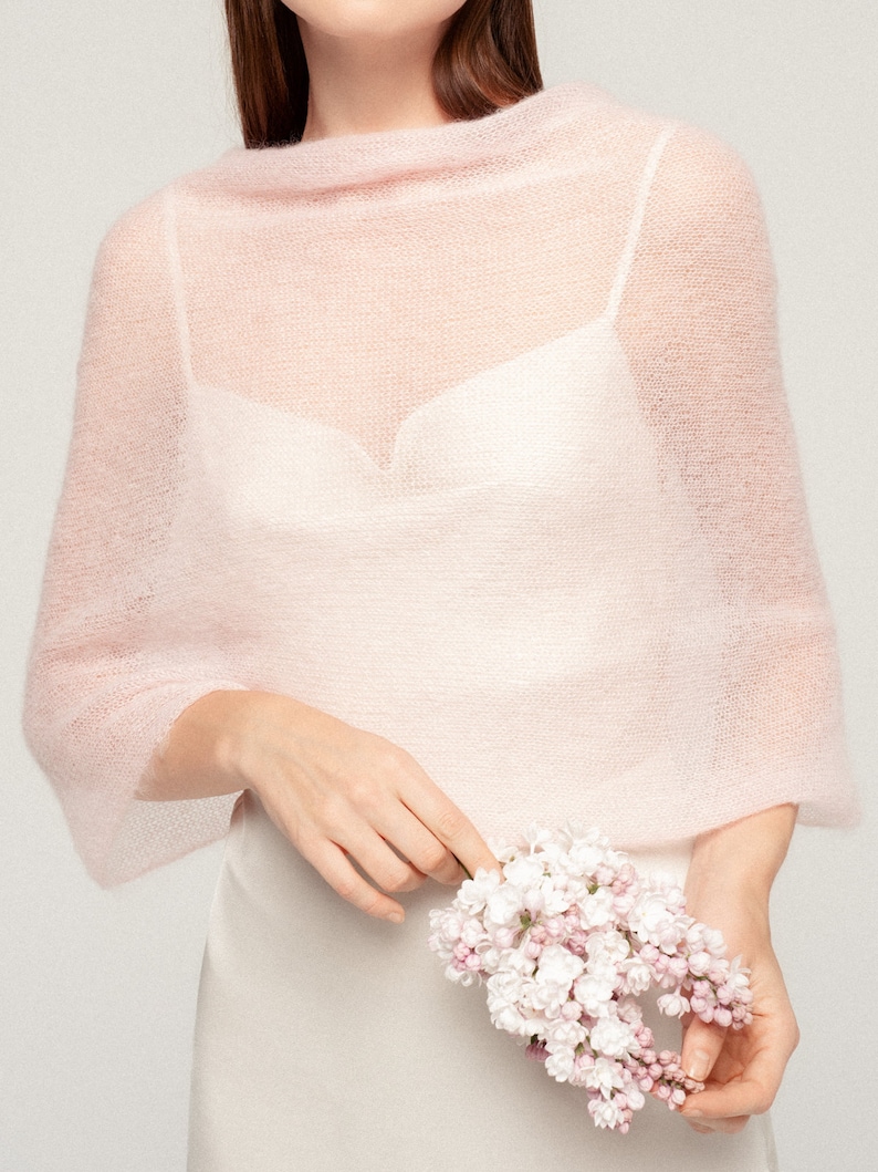 May include: A pale pink, sheer, knitted shawl with a wide boat neckline and long sleeves, worn over a white camisole. The garment features a delicate, airy texture and a loose, draped fit that covers the shoulders and upper arms.