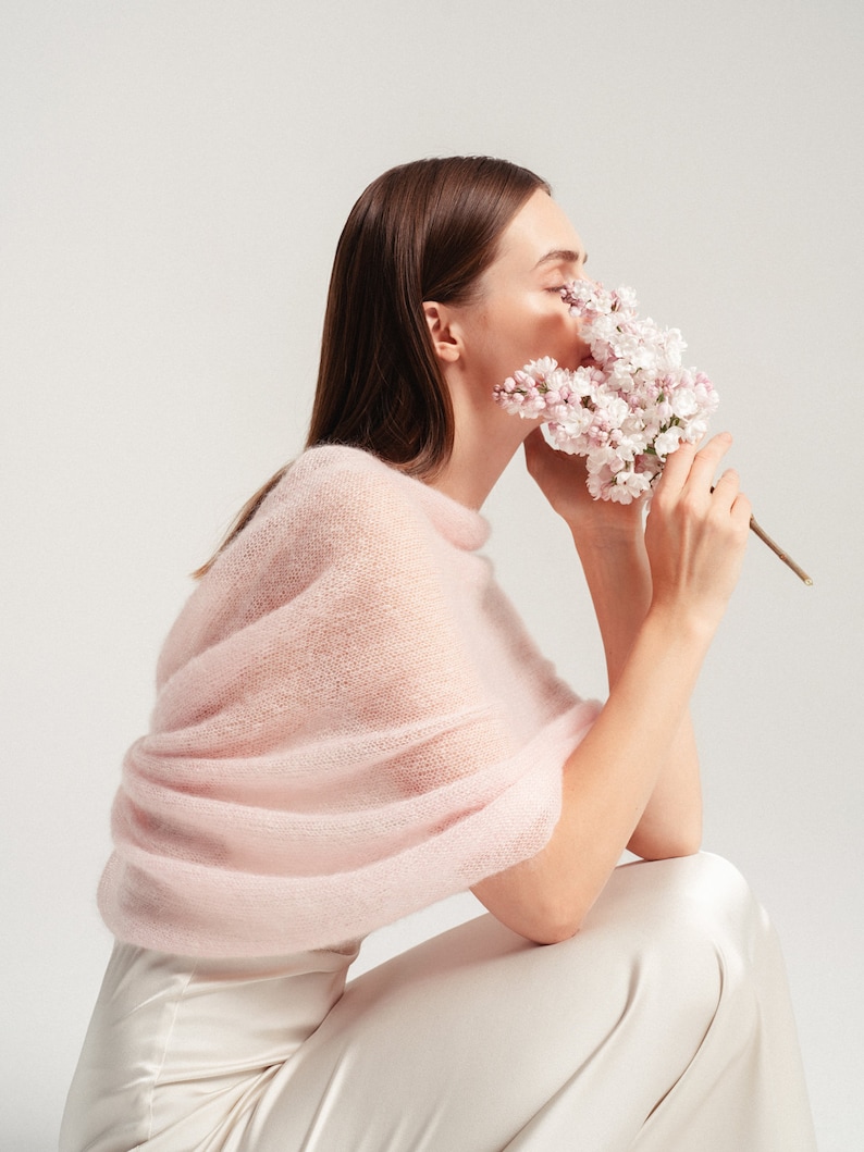 May include: A soft, pale pink knitted shawl with a fuzzy, textured finish draped over the shoulders. The garment features a wide, wrap-around design that covers the upper torso and arms, paired with smooth, cream-colored trousers.