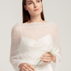 May include: A sheer white knitted bolero with a wide boat neckline and long sleeves, worn over a white camisole. The garment features a fine, semi-transparent texture and a lightweight construction that drapes softly across the shoulders and arms.