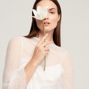 May include: A sheer, white, long-sleeved knit sweater with a fine, airy texture, worn over a solid white camisole. The garment features a simple, round neckline and a lightweight, semi-transparent construction that reveals the layer underneath.