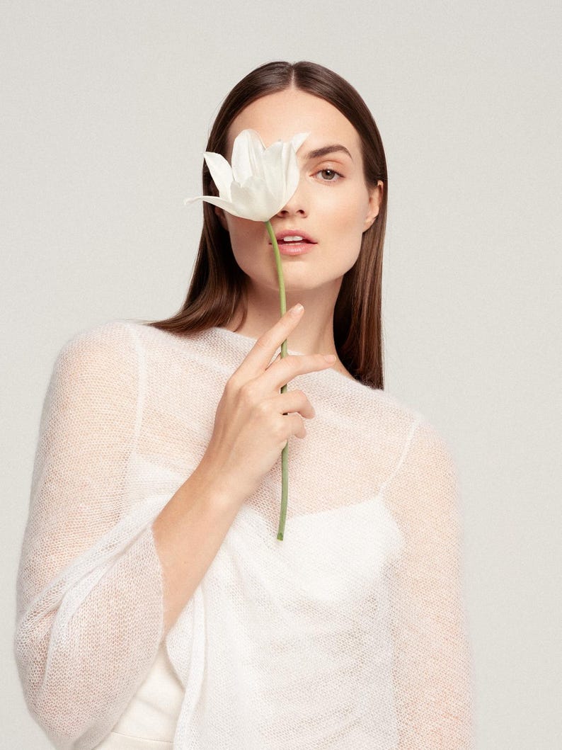 May include: A sheer, white, long-sleeved knit sweater with a fine, airy texture, worn over a solid white camisole. The garment features a simple, round neckline and a lightweight, semi-transparent construction that reveals the layer underneath.