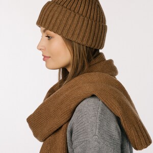 May include: A brown ribbed knit beanie with a folded cuff and a matching brown knit scarf draped over the shoulders, worn over a gray long-sleeve sweater. The set features a consistent textured knit pattern throughout the beanie and the scarf.
