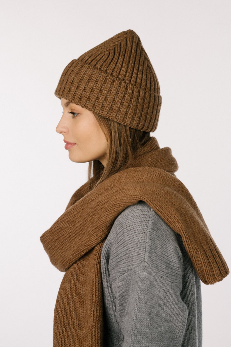 May include: A brown ribbed knit beanie with a folded cuff and a matching brown knit scarf draped over the shoulders, worn over a gray long-sleeve sweater. The set features a consistent textured knit pattern throughout the beanie and the scarf.