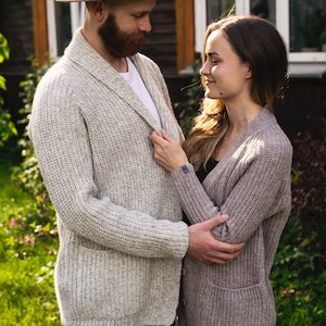 May include: A couple standing in a garden, looking at each other. The man is wearing a light brown fedora hat and a light gray cardigan. The woman is wearing a light brown cardigan.