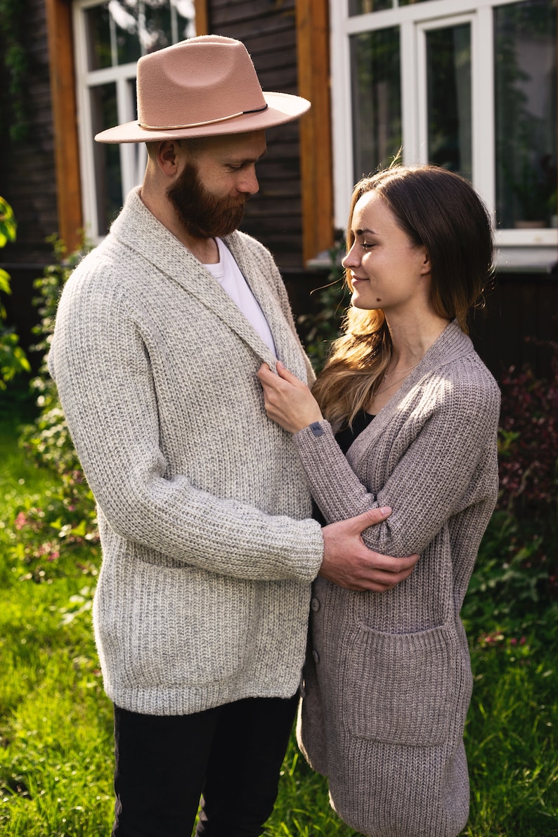 May include: A couple standing in a garden, looking at each other. The man is wearing a light brown fedora hat and a light gray cardigan. The woman is wearing a light brown cardigan.