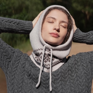 Könnte beinhalten: Graue Strickkapuze mit Kordelzug, getragen über einem passenden grauen Zopfstrickpullover. Die Kapuze hat ein Muster in Braun und Creme. Das Model hat die Arme erhoben, die Hände am Kopf und die Augen geschlossen.