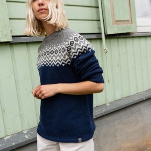 May include: A woman wearing a navy blue sweater with a grey and white geometric pattern. The sweater has a crew neck and long sleeves. She is wearing white pants.